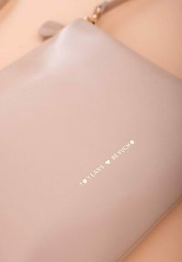 Světle růžová kožená clutch s hladkým povrchem. Na přední straně obsahuje zlatý embosovaný text "10 LET ❤️ BE VUCH". Detaily se zipem.
