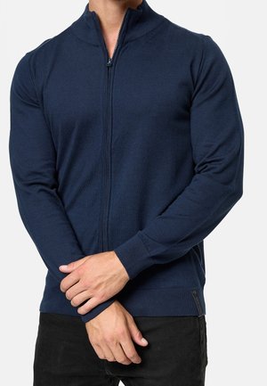 Mann trägt einen dunkelblauen Zip-Up-Pullover mit gerippten Bündchen, steht mit vor der Brust verschränkten Händen vor einem einfarbigen hellen Hintergrund.