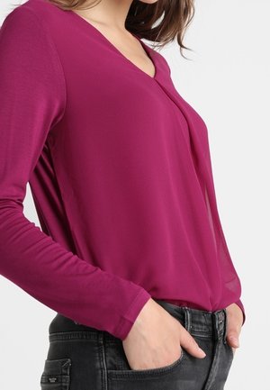 Nainen, joka käyttää vartalonmyötäistä pitkähihnaista magentanväristä paitaa, joka on tungettu korkeavyötäröisten mustien farkkujen sisään, molemmat kädet taskuissa.