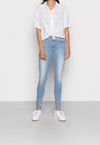 Mujer vestida con jeans ajustados de color azul claro, zapatillas blancas y una camisa blanca de manga corta abotonada atada a la cintura.