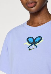 Ανοιχτό μοβ βαμβακερό μπλουζάκι με γραφικό δύο μπλε ρακέτες τένις σε φόντο κίτρινων και μπλε πιτσιλιών; λογότυπο της Nike σε πράσινο.