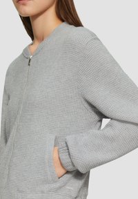 Pull en maille grise texturée avec une fermeture éclair, col rond et poches latérales. Présente un design en tricot gaufré et un matériau doux et léger.