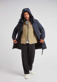 Giacca puffer blu navy con cappuccio, che mostra una giacca beige con zip sotto, foderatura interna grigia e pantaloni neri a sigaretta. Sneakers bianche in vista.