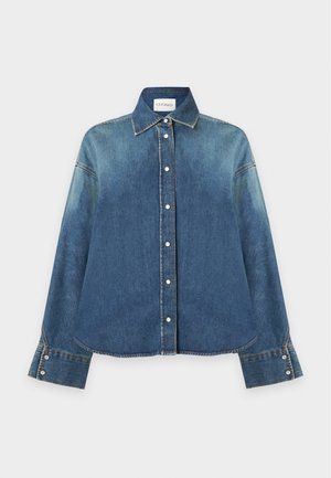 Frau, die eine blaue Jeansjacke, ein gestreiftes Hemd, zweifarbige Jeans und schwarze spitze Ballerinas trägt, steht vor einem schlichten grauen Hintergrund.