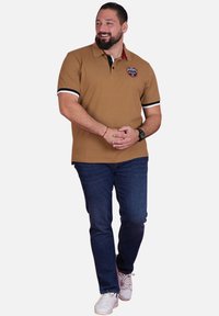 Polo marron à manches courtes, avec des accents noirs et blancs, et un logo. Associé à un jean bleu foncé et des baskets blanches.