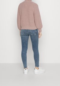 Pull tricot rose clair texturé avec un col montant, associé à un jean bleu ajusté et des baskets blanches. Vue de dos montrant la coupe de la tenue.