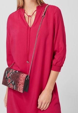 Jasnoróżowa bluzka z dekoltem w kształcie litery V i sznurowanym detalem. Modelka niesie małą czarną torebkę z czerwono-czarnym wzorem węża.