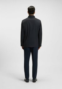 Chaqueta oscura y entallada con una textura suave, que presenta un cuello clásico y un corte recto, acompañada de pantalones azul marino y zapatos negros.