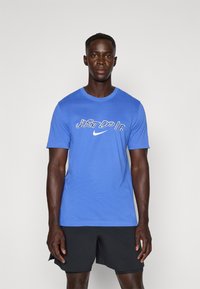 Sininen puuvillapaita, jossa on valkoiset "JUST DO IT" -teksti ja Nike-logo. Paidassa on lyhyet hihat ja klassinen pyöreä kaula-aukko.