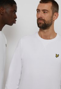 White cotton sweatshirt featuring a crew neckline and ribbed cuffs, with a small yellow embroidered logo on the chest.