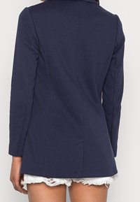 Marinblå skräddarsydd kavaj med långa ärmar, skräddarsydd design, slät tyg och detalj med sprund bak, tillsammans med slitna vita shorts.