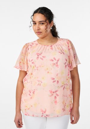 Žena s tmavými kučeravými vlasmi, oblečená v svetloružovej kvetinovej blúzke s krátkymi volánovými rukávmi a bielych nohaviciach na bielom pozadí.