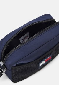 Borsa a tracolla blu navy con tasca frontale in rete nera, patch con logo Tommy Jeans e zip parzialmente aperta che mostra l'etichetta interna.