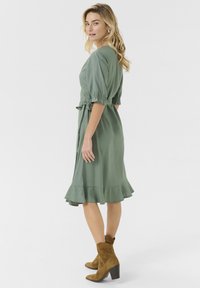 Green midi dress with puff sleeves, tie waist detail, and a ruffled hem. Styled with brown ankle boots and a smooth fabric texture.