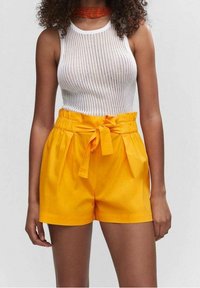 Pantaloni corti gialli in cotone con vita a sacco di carta, dotati di una cintura legata e tasche laterali. Abbinati a un top aderente bianco a coste.