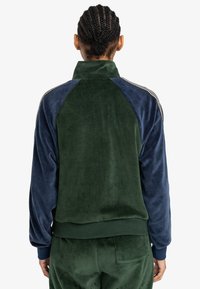 Chaqueta de chándal de terciopelo con cuello alto, cuerpo verde y mangas azul marino, puños acanalados y acentos laterales en contraste.