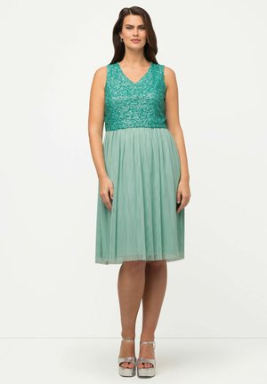 Knielanges Kleid mit einem türkisfarbenen Pailletten-Oberteil und einem fließenden mintgrünen Rock. Besitzt einen V-Ausschnitt und einen transparenten Saum. Kombiniert mit silberfarbenen Plateau-Heels.
