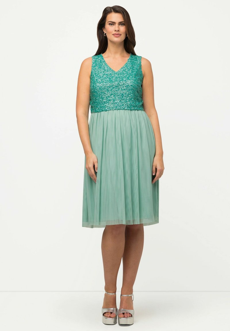 Abito al ginocchio con un corpetto di paillettes color teal e una gonna fluida verde menta. Presenta una scollatura a V e orlo trasparente. Abbinato a tacchi a piattaforma argentati.