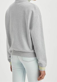 Sudadera gris de forro polar con cuello alto, puños acanalados con rayas amarillas y un corte holgado. Combinada con jeans azul claro.