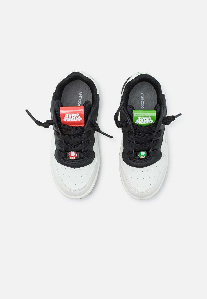 Baskets blanches et noires avec des lacets noirs, avec des étiquettes de langue Super Mario rouges et vertes et des icônes de champignons sur les lacets.