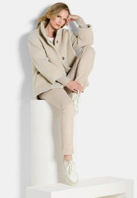 Beige textuurjasje met een sjawlkrage en twee knopen, gecombineerd met lichtbeige broek en witte sneakers, zittend op een wit voetstuk.