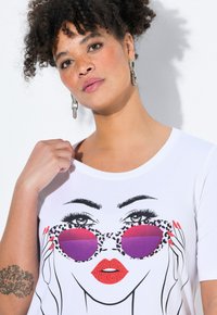 T-shirt blanc présentant un graphique d'un visage avec des lunettes de soleil imprimé léopard, des verres teintés violets, des lèvres rouges, et des cheveux et yeux dessinés en noir.