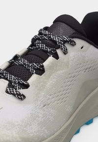 Κοντινό πλάνο ενός αθλητικού παπουτσιού ανοιχτού γκρι χρώματος με κορδόνια σε ασπρόμαυρο μοτίβο, μαύρη επένδυση στη γλώσσα και υφασμάτινο επάνω μέρος με υφή.