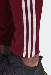 Maroon joggers with white triple stripes along the side, made of textured fabric, paired with dark athletic shoes featuring a rugged sole.