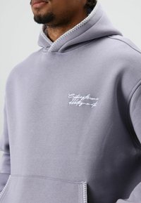Lichtpaarse hoodie met witte stiksels, voorzien van een voorzak en geborduurde tekst op de borst. Zachte stof met een relaxte pasvorm.