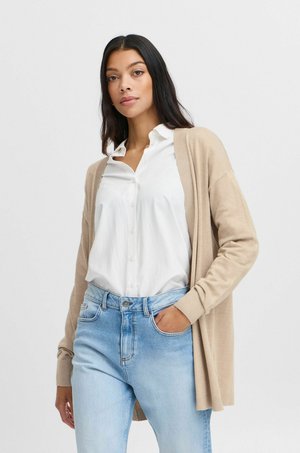 Beige Strickjacke mit langen Ärmeln über einem weißen Hemd, kombiniert mit hellblauen Jeans. Schlicht, entspannte Passform, weiche Textur sichtbar.