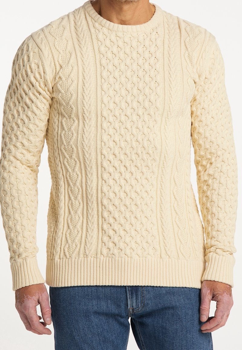 Cremfarbener Strickpullover mit strukturierten Zopfmustern, gerippten Bündchen und Saum, Rundhalsausschnitt. Kombiniert mit blauen Jeans.