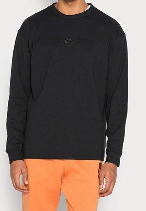 Personne portant un haut à manches longues noir de la marque Nike et un pantalon orange Nike, debout devant un fond uni clair.
