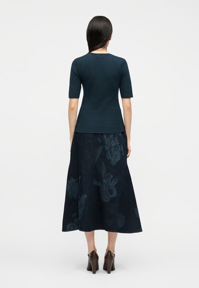 Γυναίκα με μακριά σκουρόχρωμα μαλλιά φοράει εφαρμοστή σκουροτιρκουάζ μπλούζα, μια μέση-μήκους φούστα με λουλούδια σε ναυτικό μπλε και καφέ ψηλοτάκουνα παπούτσια, εμφανιζόμενη από πίσω.