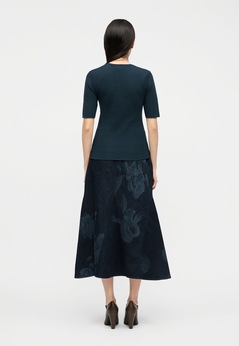 Donna con capelli lunghi e scuri che indossa una camicia aderente color verde teal scuro, una gonna midi floreale blu navy e scarpe con tacco marroni, vista da dietro.