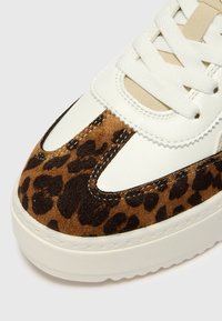 Gros plan de l'avant de la basket en cuir blanc, accents en tissu beige, lacets blancs et détail en daim marron à motif léopard.
