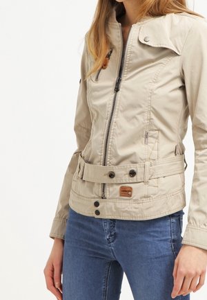 Žena v béžové bundě na zip s pásem a modrých džínách, stojící s uvolněnýma rukama podél těla.