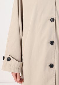 Manteau trench beige à coupe ample, avec des boutons noirs et un détail de poignet. Fabriqué dans un tissu lisse avec une texture subtile.