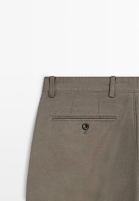 Tył szarych spodni z paskiem w talii, szlufkami na pasek oraz pojedynczą kieszenią z patką zapinaną na guzik na środku.