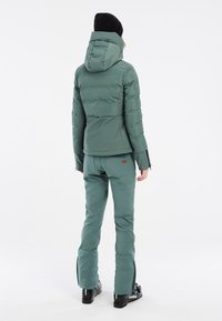 Groene geïsoleerde skijack met capuchon, gecombineerd met bijpassende skibroek. Gladde textuur, praktische ritsen en versterkte manchetten.