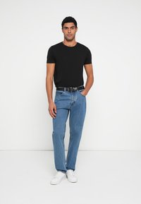 Camiseta negra de manga corta, jeans de mezclilla azul, cinturón negro y zapatillas blancas. El modelo está de pie sobre un fondo neutro.