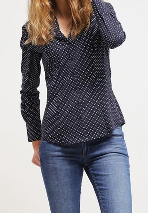 Mujer con una camisa azul marino de manga larga con lunares y jeans azules, posando con una mano tocándose el cabello.