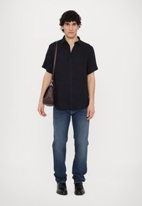 Camicetta nera a maniche corte con bottoni, jeans di denim blu, borsa in pelle marrone, scarpe nere, in piedi davanti a uno sfondo bianco.