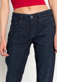 Mörkblå denimjeans med medelhög midja, framfickor och knappstängning. Tyget har en mjuk textur och subtila sömdetaljer.