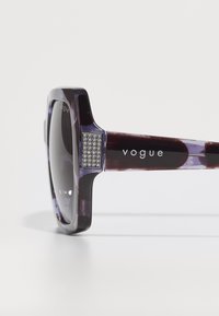 Ochelari de soare Vogue cu o formă pătrată mare, cadru violet translucid, lentile gri și un accent încrustat cu cristal pe templu.