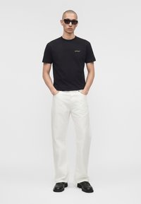 Homme portant des lunettes de soleil noires, un t-shirt noir à manches courtes avec un petit logo, un pantalon blanc droit et des chaussures noires, debout les mains dans les poches.
