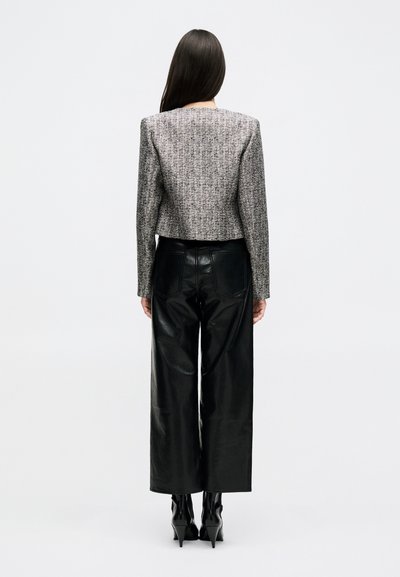 Žena s dlouhými tmavými vlasy nosí šedý krátký tweedový blejzr, černé kožené kalhoty s vysokým pasem a širokými nohavicemi a černé boty na podpatku, otočená zády.
