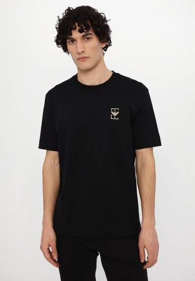 Juodos medvilnės marškinėliai trumpomis rankovėmis su mažo smėlio spalvos logotipu kairėje krūtinės pusėje ir klasikiniu apvaliu iškirpimu.