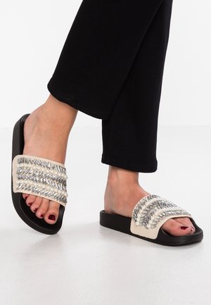 Fødder iført sorte slippers med hvide remme dekoreret med perler og sølvkugler, parret med sorte bukser.