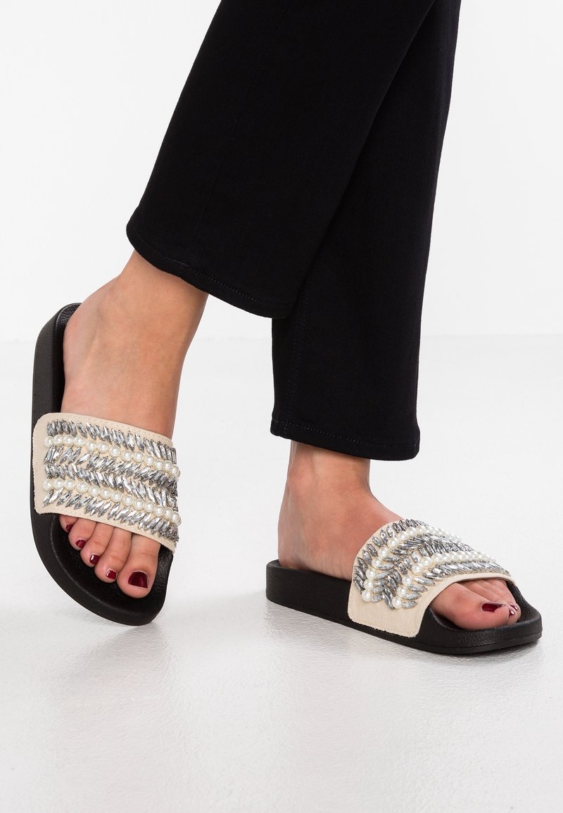 Pieds portant des sandales slides noires avec des bandes blanches décorées de perles et de perles argentées, associées à un pantalon noir.