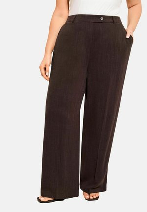 Frau trägt dunkelbraune, weit geschnittene Hosen mit Taschen, steht barfuß, eine Hand in der Tasche, die andere locker an der Seite.